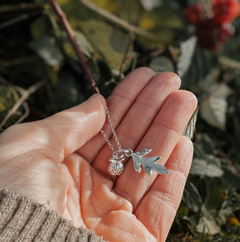 Hand holding a silver necklace with leaf and berry charms against a natural background