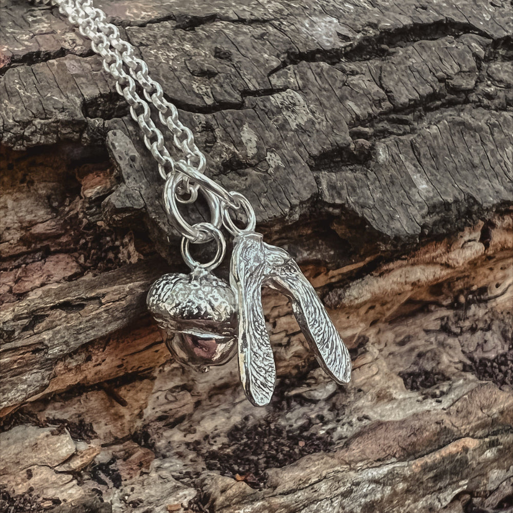 Silver necklace with acorn and sycamore seed pendant on a bark surface