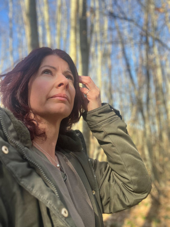 Lady walking in countryside wearing green coat and silver acorn pendant. Looking up at trees