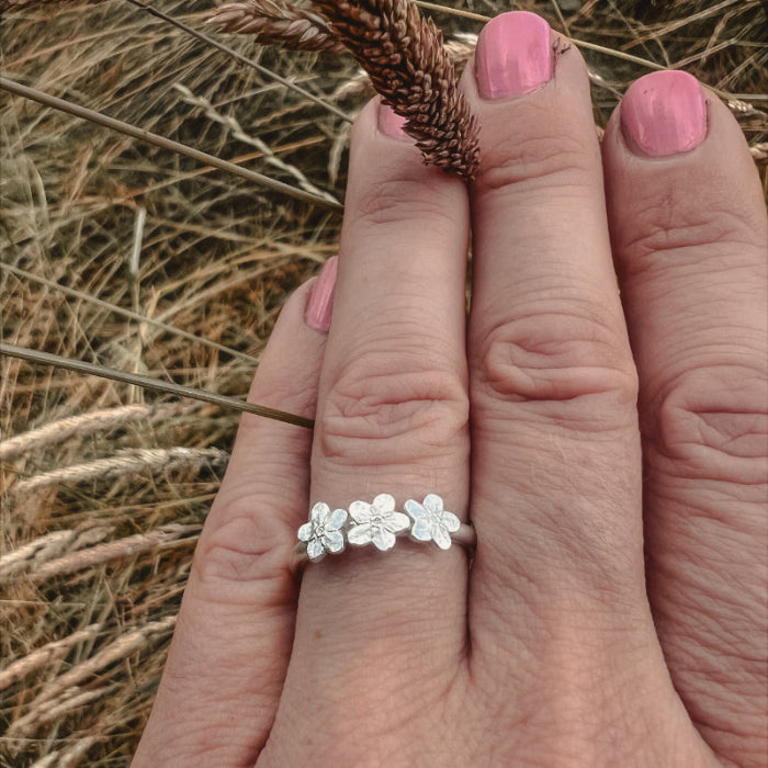 Hand with pink nails, wearing silver ring with 3 forgetmetnots with grass background