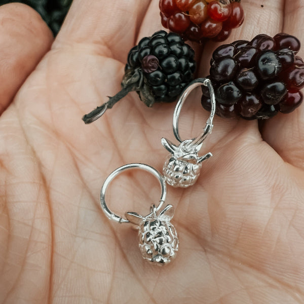 Hand with silver blackberry earrings on next to real blackberries.