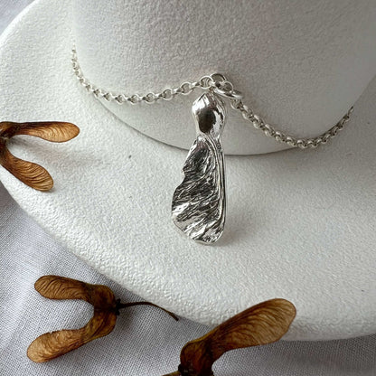 Silver seed shaped pendant on a chain with dried seeds on a white surface