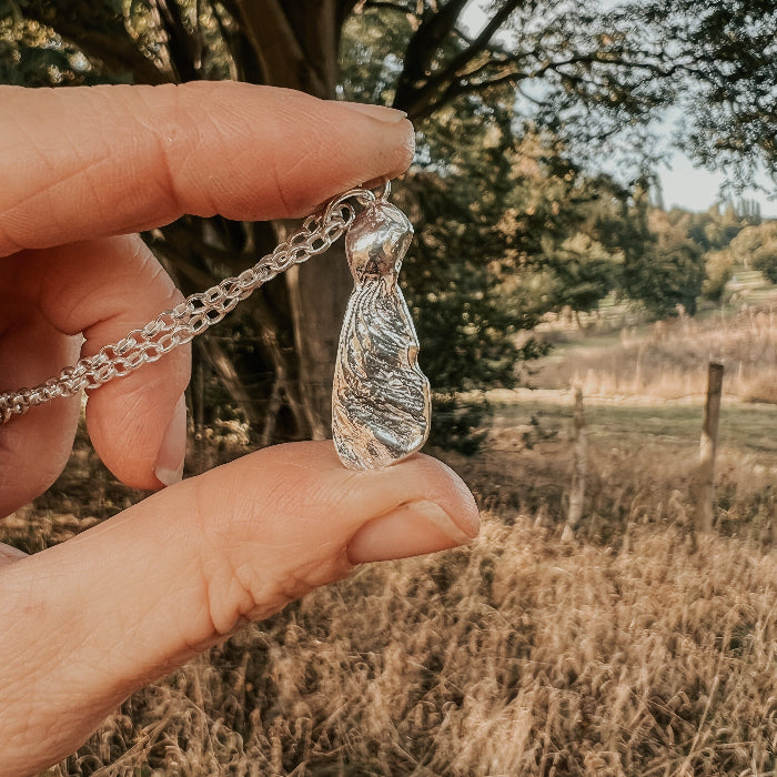 Hand holding a silver necklace with a sycamore seed pendant against a natural background