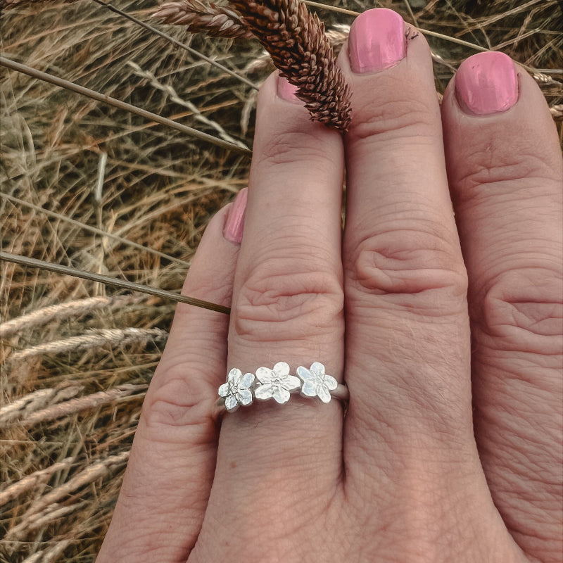 Hand with pink nails, wearing silver ring with 3 forgetmetnots with grass background